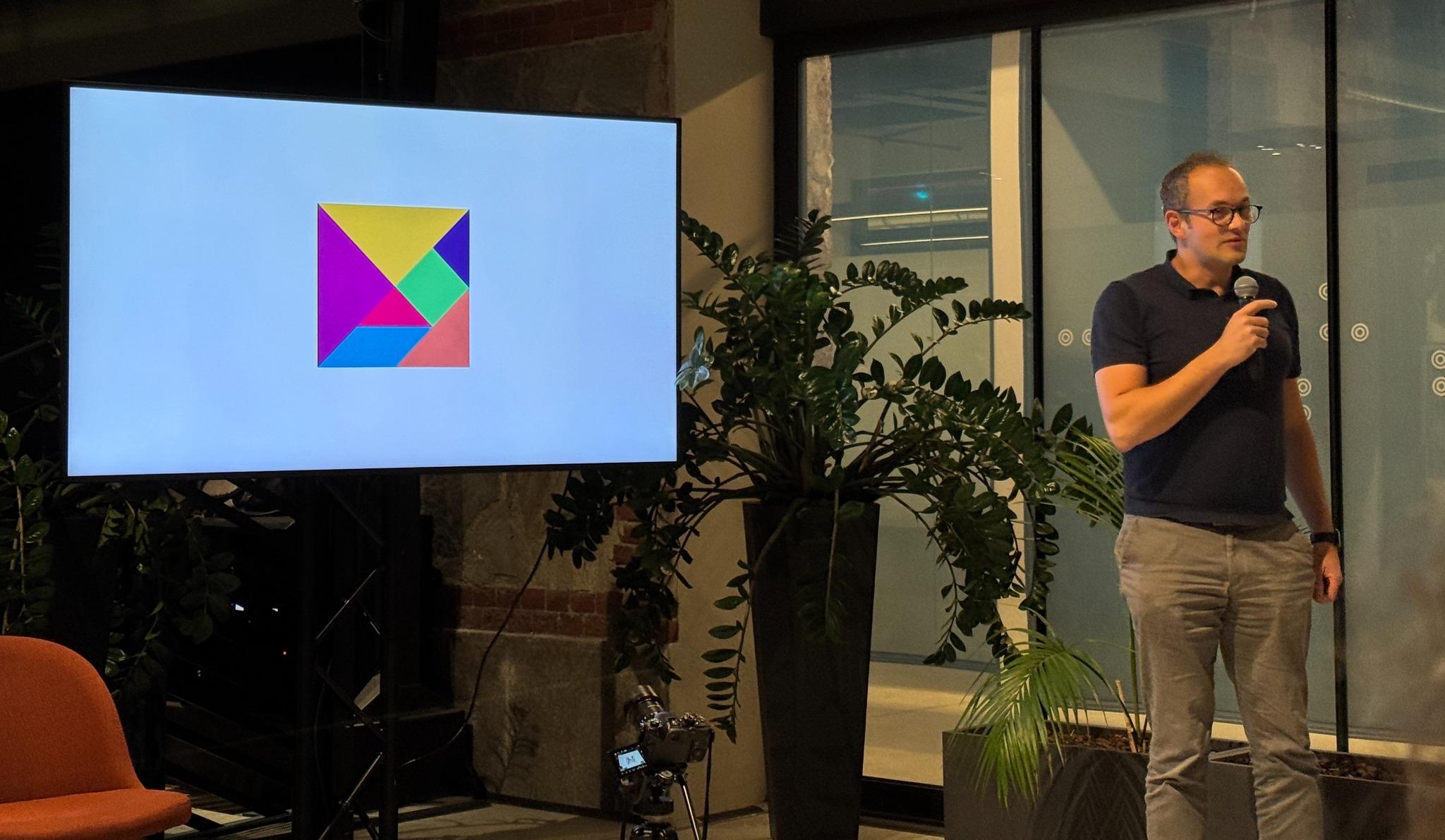 A presenter holding a microphone beside a screen showing a colourful geometric graphic at the Mindstone AI meetup in Turin.